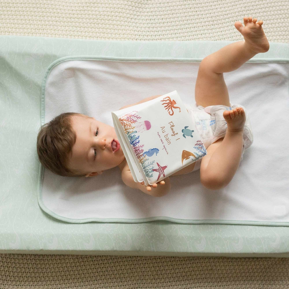 Bébé qui joue avec son livre de bain allongé en couche sur son matelas à langer